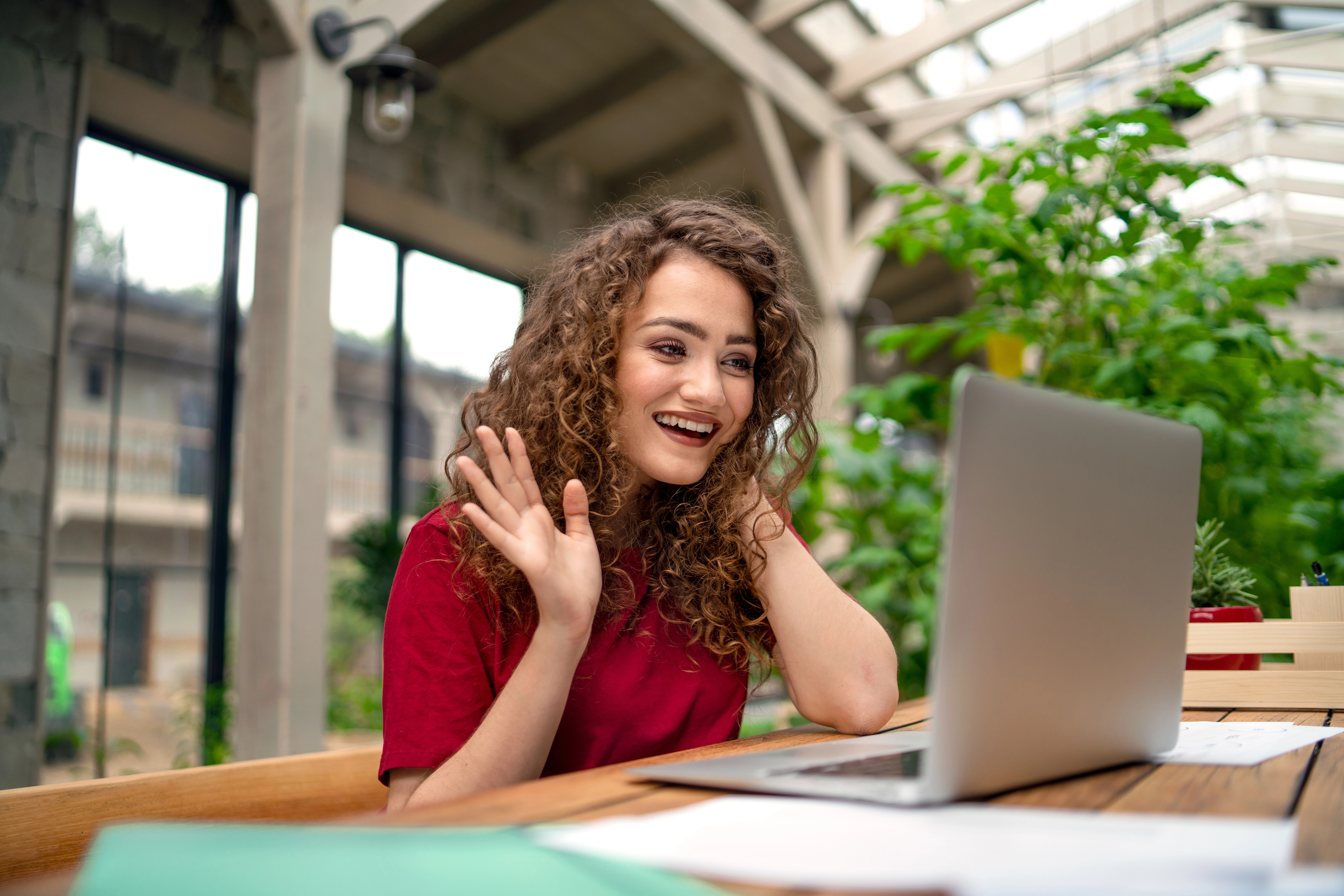 Photo of a happy smiling woman waving at a laptop, sitting in a cafe outside
