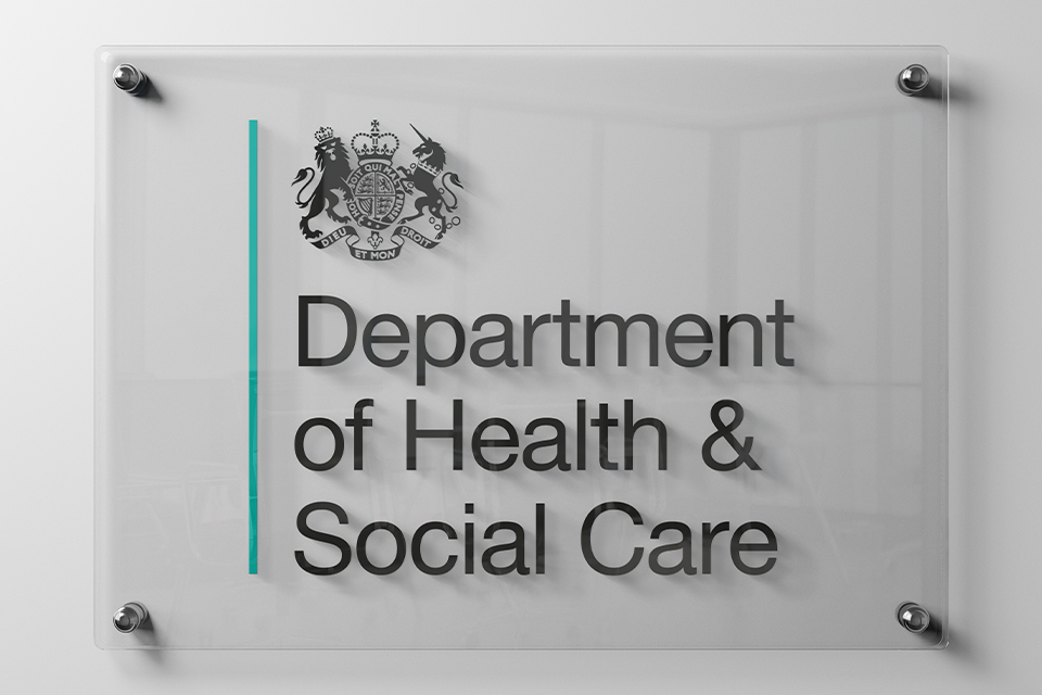 Glass sign displaying the UK Government crest and the words “Department of Health & Social Care” in large black lettering. The transparent sign is mounted on a light grey wall using four metal fixings, with a vertical turquoise line running down the left side.