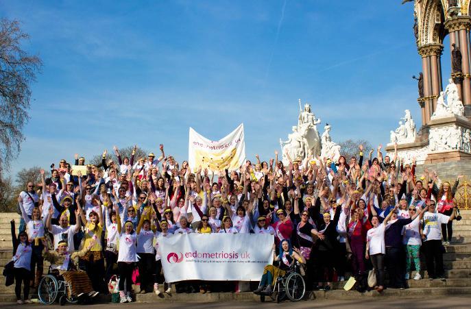 A large crowd of people gathered on the steps of the Albert Memorial in London for an "EndoMarch" event, holding up an "Endometriosis UK" banner and raising their arms in celebration. A yellow banner is held aloft in the background, and the ornate gothic memorial with its statues rises behind the group under a clear blue sky.
