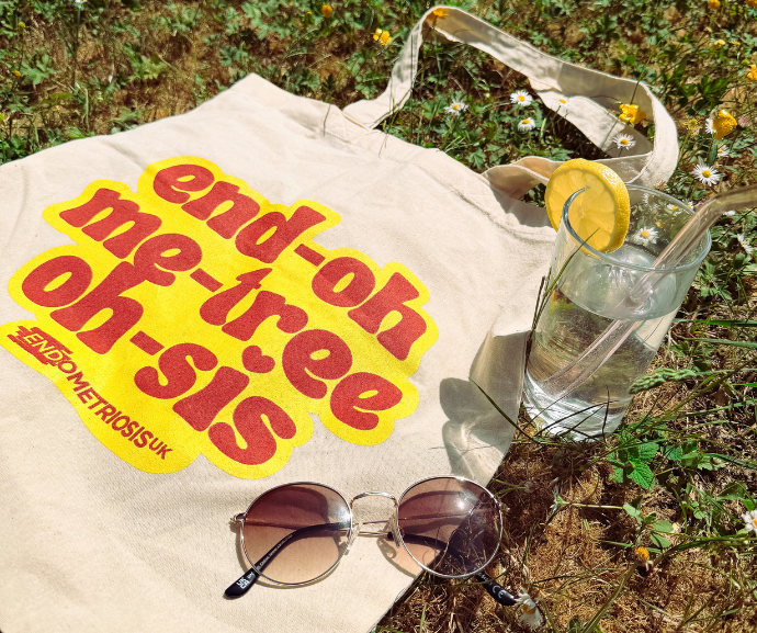 A cream tote bag lies on grass among daisies, printed with "end-oh me-tree oh-sis" in bold red bubble letters on a yellow background, spelling out "endometriosis," with "Endometriosis UK" printed below. Round tortoiseshell sunglasses rest on the bag, and beside it sits a glass of water with a lemon slice and straw.
