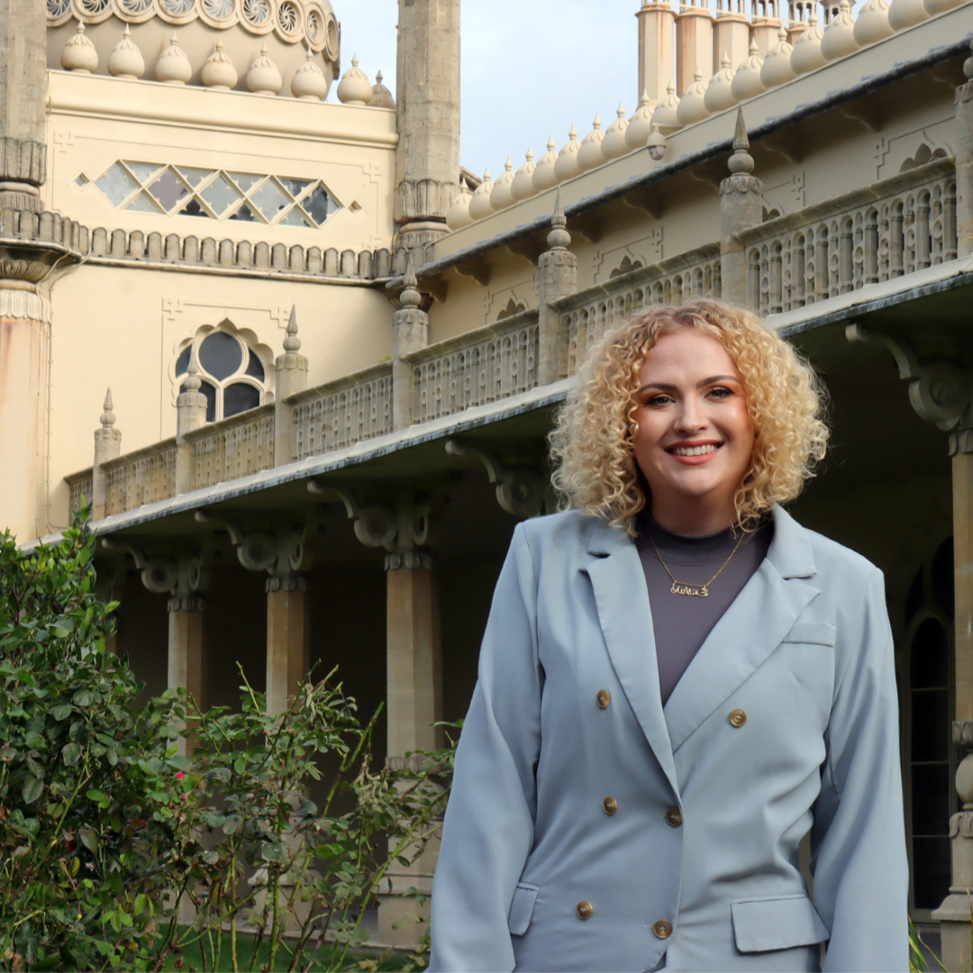 Portrait of a person standing outdoors in front of an ornate historic building with decorative arches, columns and domed architectural details. The person is wearing a light blue double-breasted blazer over a dark top and a necklace, and is positioned in a landscaped garden area with shrubs in the foreground. The building fills much of the background, creating a formal setting