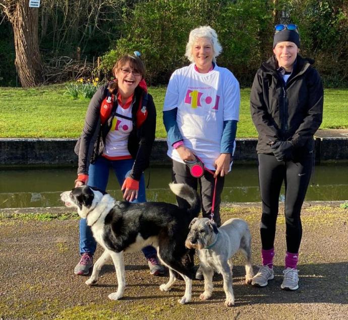 Three smiling women stand together beside a canal on a sunny day, two of them wearing white "Endometriosis UK" branded T-shirts, accompanied by two dogs, one black-and-white and one gray, on the paved towpath in front of them. Greenery and daffodils line the canal bank behind the group.