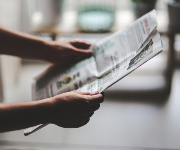Close-up photograph of a person holding and reading an open newspaper indoors. The newspaper is slightly out of focus, with hands visible supporting the pages. The background is softly blurred and includes a tabletop and household objects, creating a neutral indoor setting.