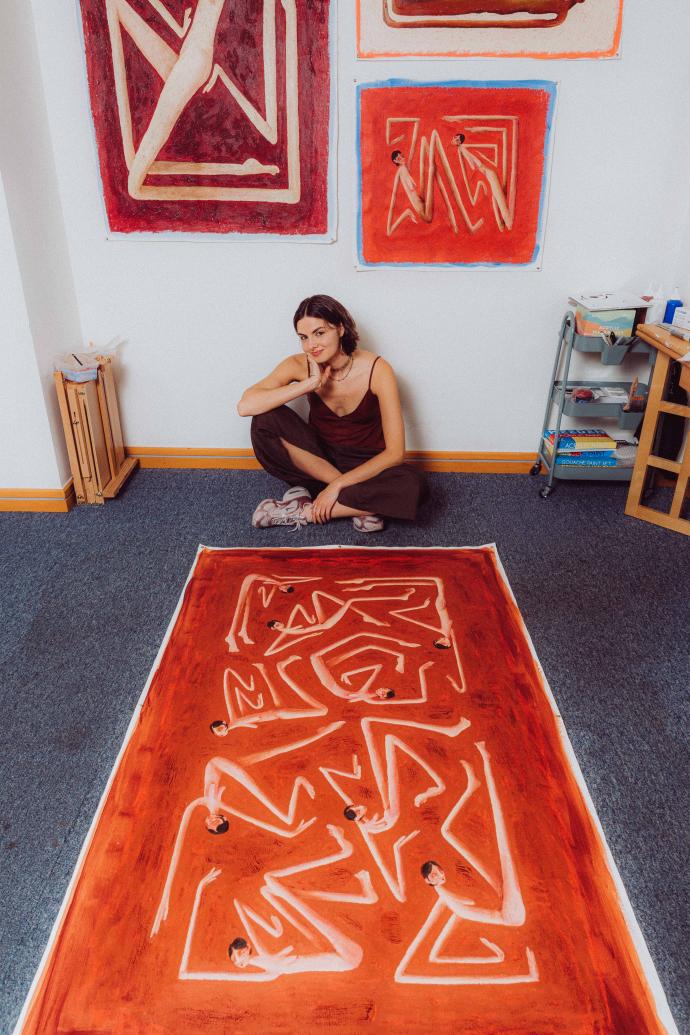 A person sits cross-legged on the floor of an art studio beside a large orange and white abstract painted rug with geometric, maze-like patterns. Behind them, several framed abstract artworks with dark angular brushstrokes hang on the wall, along with shelving holding rolled materials and supplies