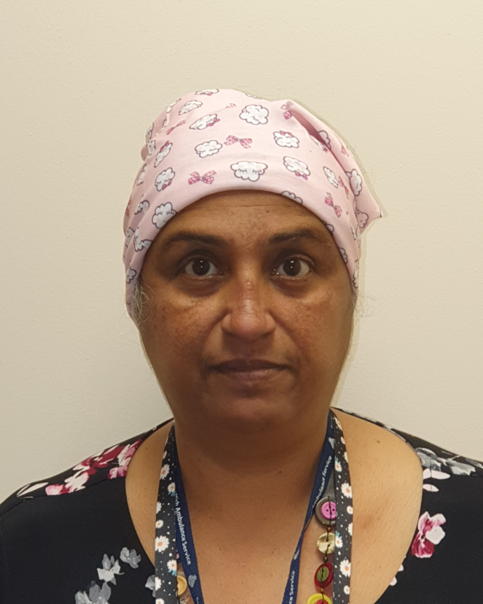 Portrait photo of Shy, a woman wearing a pink patterned headscarf and a floral top, with an Ambulance Service lanyard around her neck, looking directly at the camera against a plain wall background