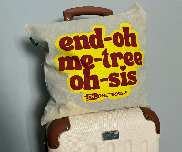 A cream tote rests on a suitcase, printed in bold red and yellow text with the phonetic spelling 'end-oh me-tree oh-sis' and the Endometriosis UK logo below it.