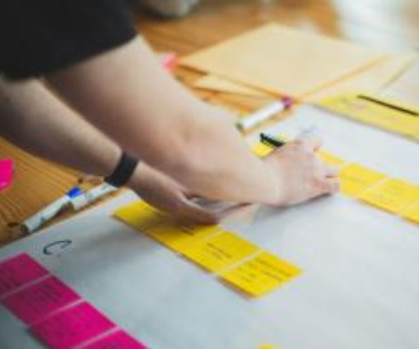 Close-up of a collaborative planning or workshop session around a tabletop covered with large sheets of paper and colourful sticky notes. A person wearing a dark top and wristwatch is placing or arranging yellow sticky notes on the table, while pink sticky notes, pens and markers are visible nearby