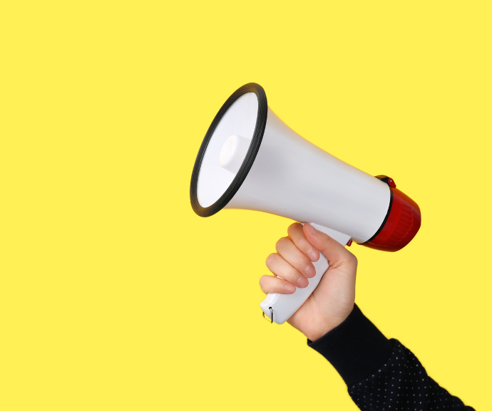 Close-up photograph of a hand holding a white and red megaphone against a solid pale yellow background. The megaphone is angled diagonally upward, with the speaker cone facing left and the handle gripped firmly. A dark sleeve is visible at the bottom right of the image.