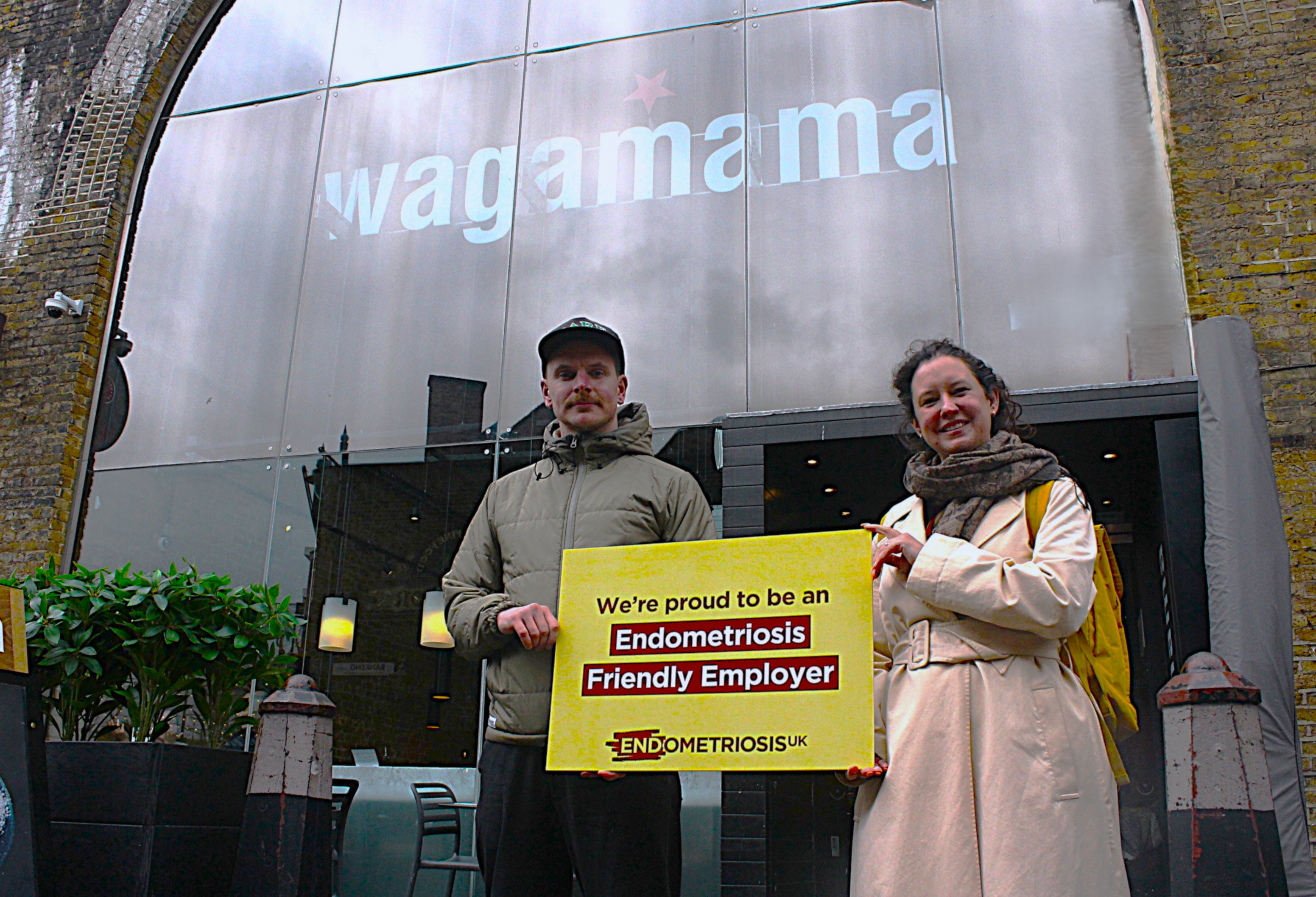 Two people stand outside a Wagamama restaurant, positioned in front of a large glass façade with the Wagamama logo displayed above the entrance. One person is wearing a light-coloured padded jacket and cap, while the other is wearing a light trench coat and scarf with a yellow backpack. Together they hold a large yellow sign that reads: “We’re proud to be an Endometriosis Friendly Employer”, with the Endometriosis UK logo beneath. The restaurant entrance, outdoor seating and planters are visible in the fore