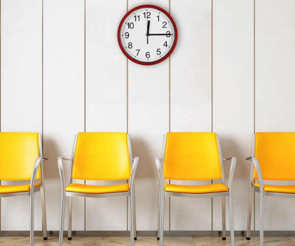 Waiting room with four bright yellow chairs arranged against a light-coloured panelled wall. A round wall clock with a red frame hangs above the chairs.