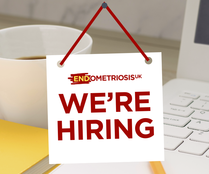 A hanging sign with the Endometriosis UK logo reads 'We're Hiring' in bold red text, displayed against a background of a coffee cup, laptop keyboard, and notebook.