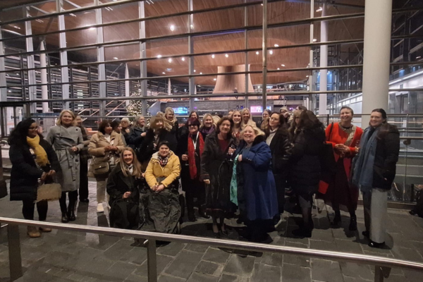 Group photograph of around 20 people gathered inside a large modern building with floor-to-ceiling glass walls and a high wooden ceiling. Several people are standing while others are seated or using mobility aids in the front row. The group is arranged for a photo on a dark stone floor behind a metal handrail.