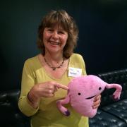 A smiling woman with short brown hair, wearing a yellow top and a name badge, holds and points to a pink plush toy shaped like a uterus, while seated on a black leather sofa.
