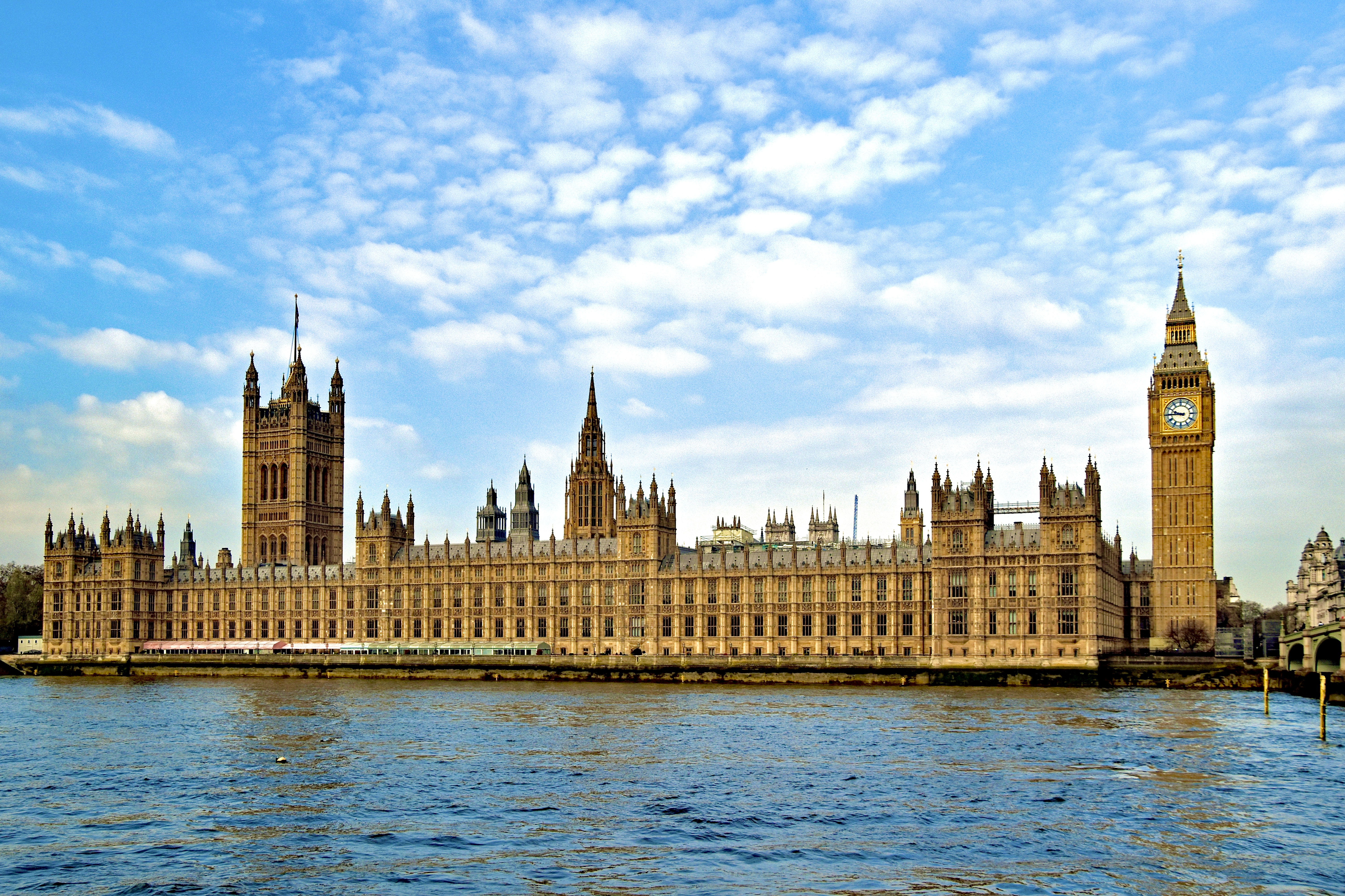 Wide view of the Palace of Westminster beside the River Thames in London. The historic Gothic Revival building stretches across the image, with the Elizabeth Tower, commonly known as Big Ben, standing prominently on the right. The River Thames occupies the foreground, while a bright blue sky with scattered white clouds fills the background.