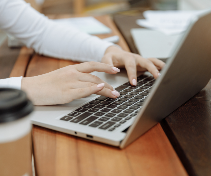 Close-up photograph of hands typing on a laptop computer placed on a wooden table. The laptop keyboard and trackpad are in focus, while a takeaway coffee cup is partially visible in the foreground and papers and office items are softly blurred in the background.