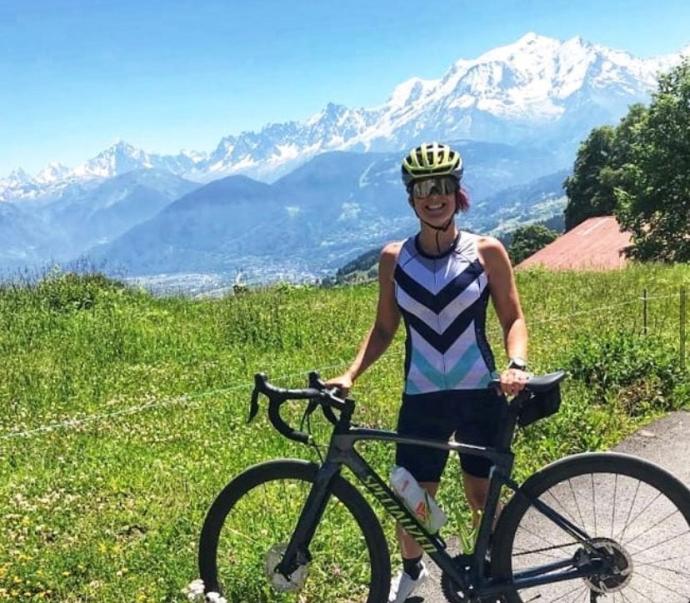 A woman in a striped cycling outfit, helmet, and sunglasses stands smiling with her road bike on a grassy hillside, with dramatic snow-capped mountains and a valley stretching out behind her under a clear blue sky.