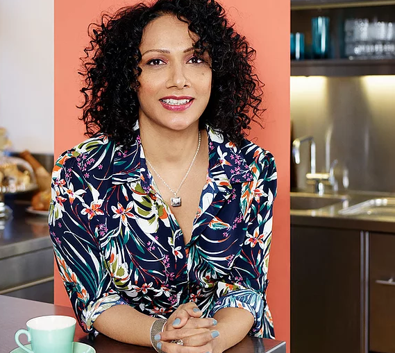 Portrait of a person with dark curly hair seated at a kitchen counter, wearing a navy shirt with a colorful floral pattern and a silver necklace. Hands are clasped on the countertop beside a pale green cup. The background includes a peach-colored wall panel and a modern kitchen with shelves, cabinets, and a sink.