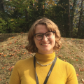 A woman with short, curly brown hair and glasses smiles at the camera, wearing a mustard-yellow top and a lanyard, standing outdoors with autumn trees in the background.