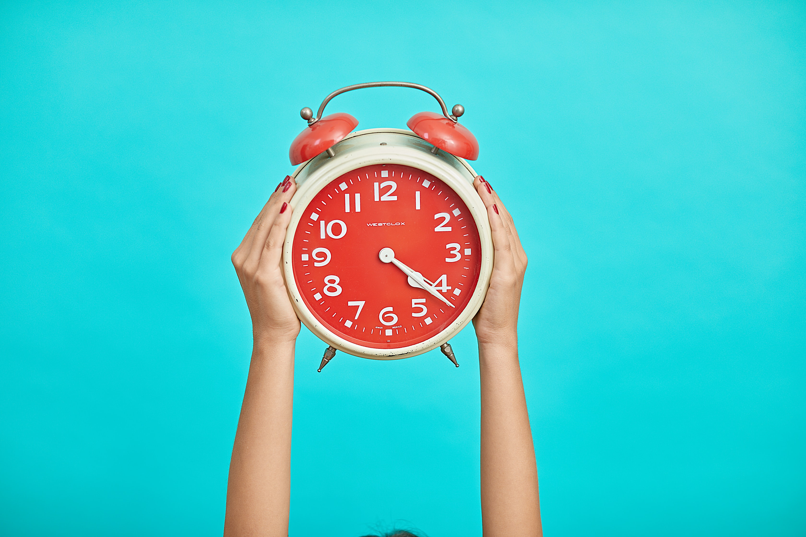 Person holding a large vintage-style red alarm clock above their head against a bright turquoise background. The clock face has white numbers and hands, while the person's hands, with red-painted fingernails, grip each side of the clock.