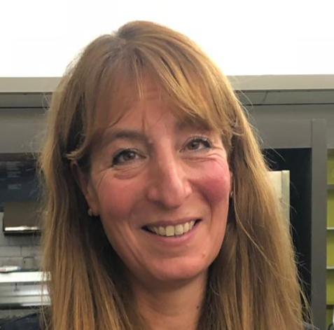 A close-up portrait of a smiling woman with long, straight blonde hair and bangs, photographed indoors.