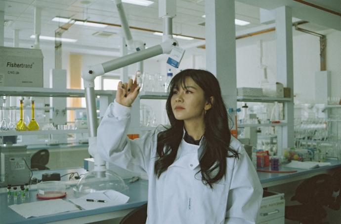 A woman in a white lab coat stands in a laboratory, holding up a small glass vial and examining it closely. She's surrounded by lab equipment, including a Fisherbrand-labeled shelf, glass flasks with yellow liquid, an articulated overhead lamp or arm, and various bottles and petri dishes on the counter in front of her.