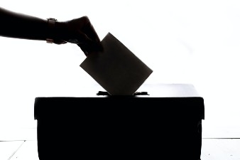 Silhouette of a hand placing a ballot paper into a ballot box against a bright white background.