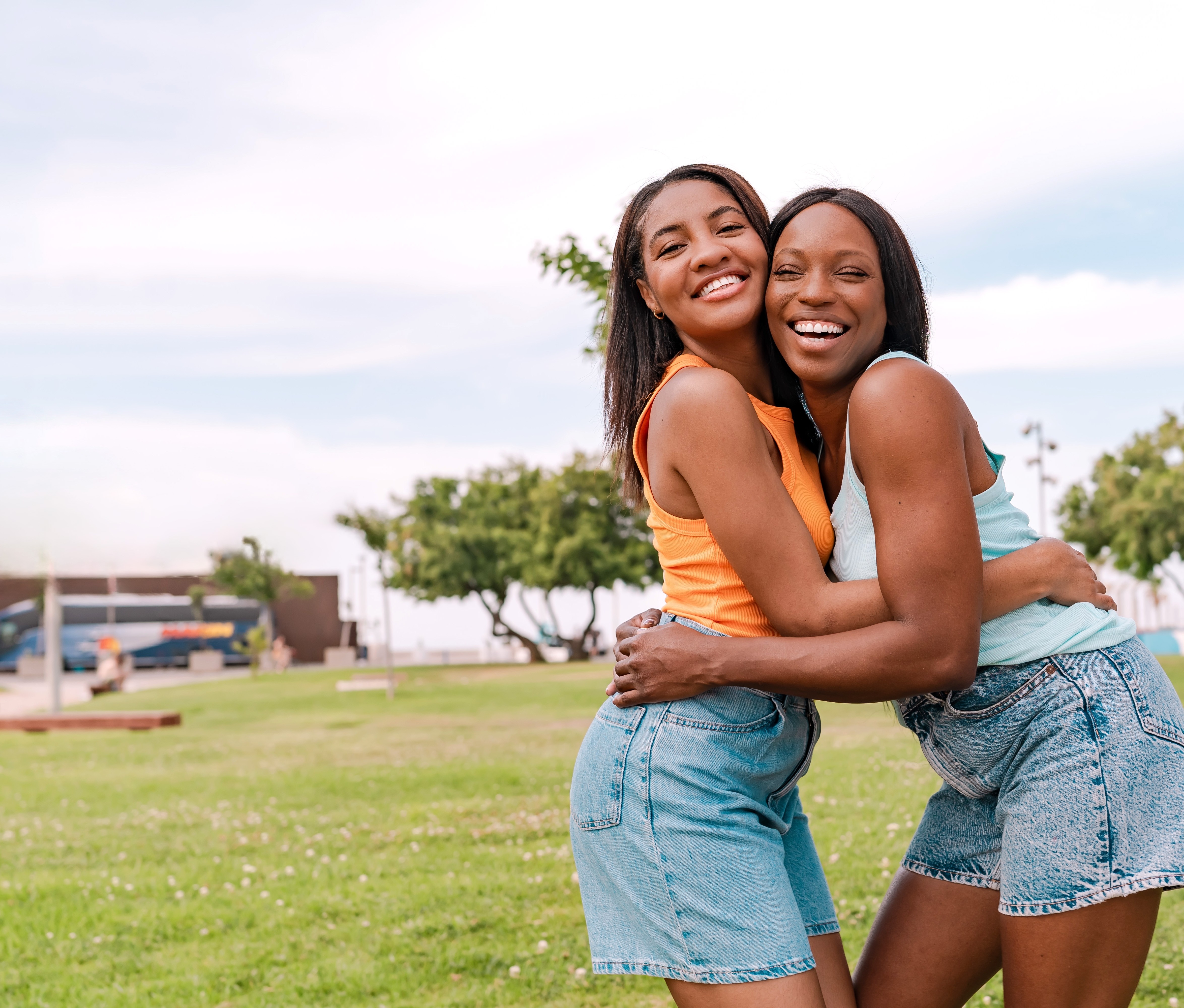 Two people standing on a grassy field outdoors with trees and an overcast sky in the background. The pair are embracing and facing the camera, wearing sleeveless tops and denim shorts. The open park setting includes a lawn stretching across the foreground, with scattered trees, a pathway and distant buildings visible in the background.