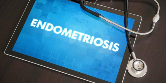 A tablet screen displays the word "ENDOMETRIOSIS" in bold white text over a blue medical-themed background with a heartbeat line graphic, with a stethoscope draped across the tablet on a dark wooden surface.