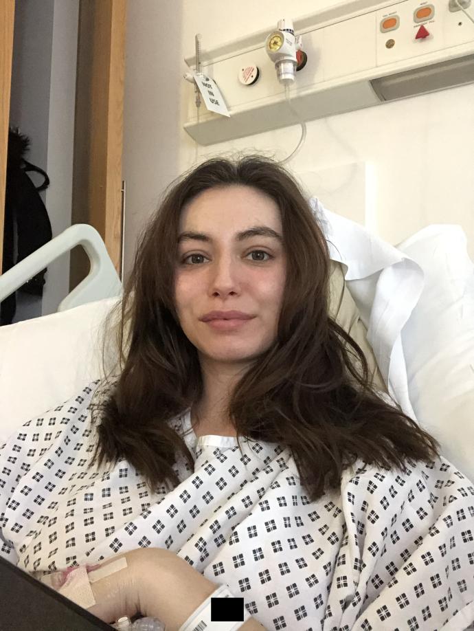 A woman with long, wavy brown hair sits up in a hospital bed, wearing a patterned hospital gown, looking toward the camera with a slight smile. A hospital ID wristband and a bandage on her arm are visible, and medical equipment, including oxygen and call buttons, is mounted on the wall behind her.