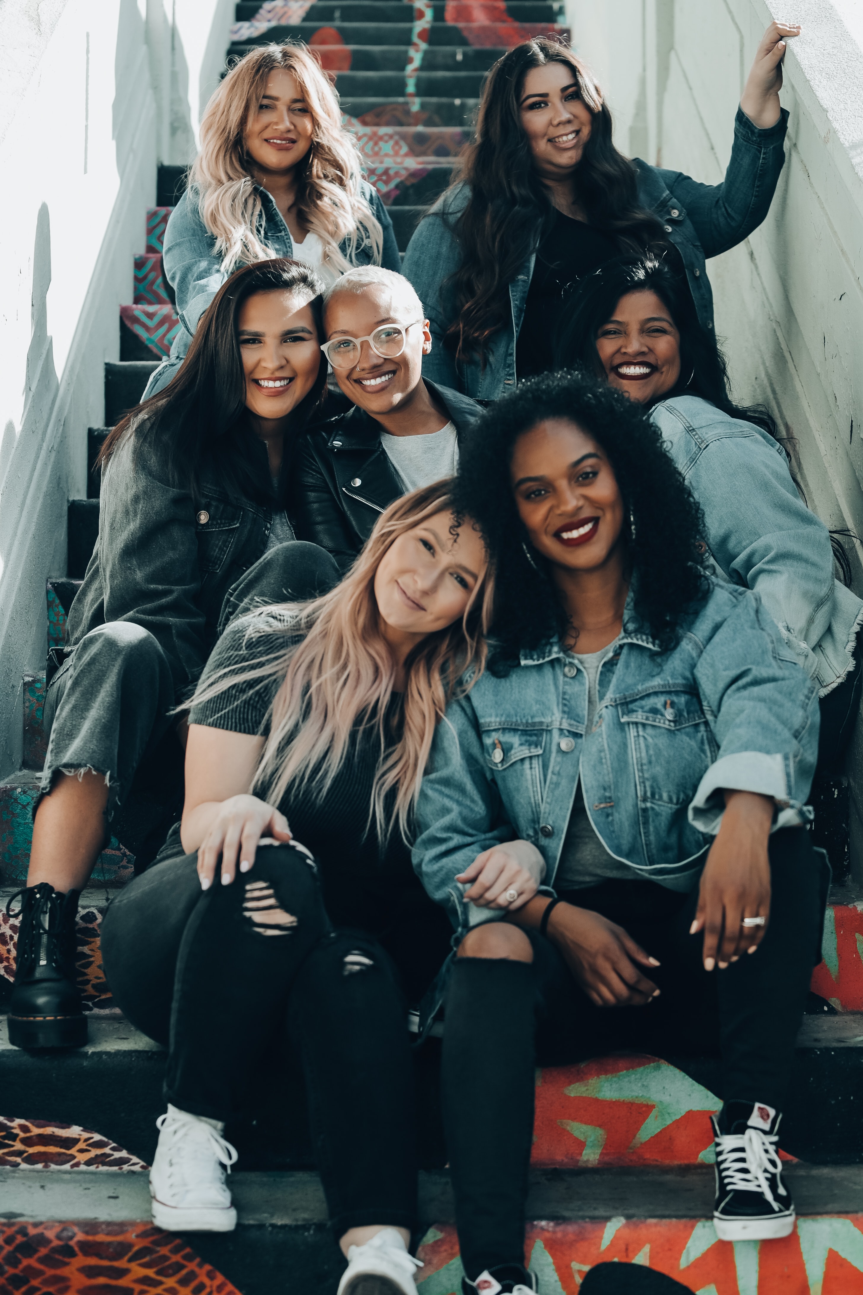 Group portrait of seven people seated and standing on a painted staircase indoors. The individuals are arranged across several steps and wear casual clothing, including denim jackets, dark tops and jeans. The staircase features colourful patterned artwork, while natural light from the side illuminates the scene