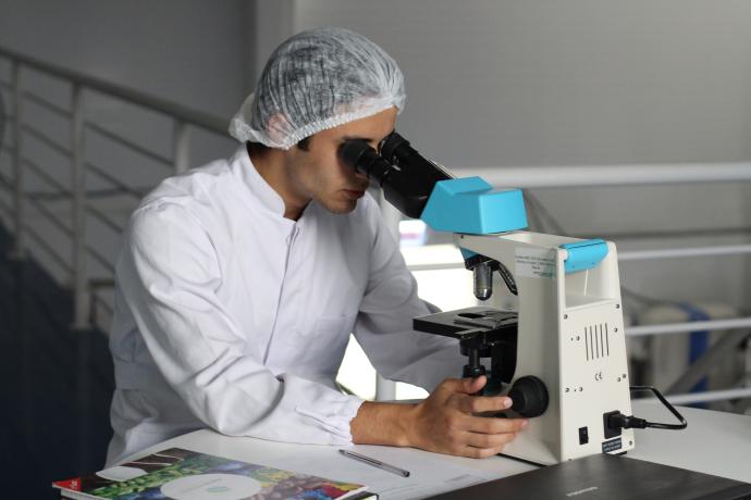 A man wearing a white lab coat and a hairnet leans over a table, looking through the eyepiece of a blue and white microscope. Papers, pens, and a laptop are visible on the desk in front of him.