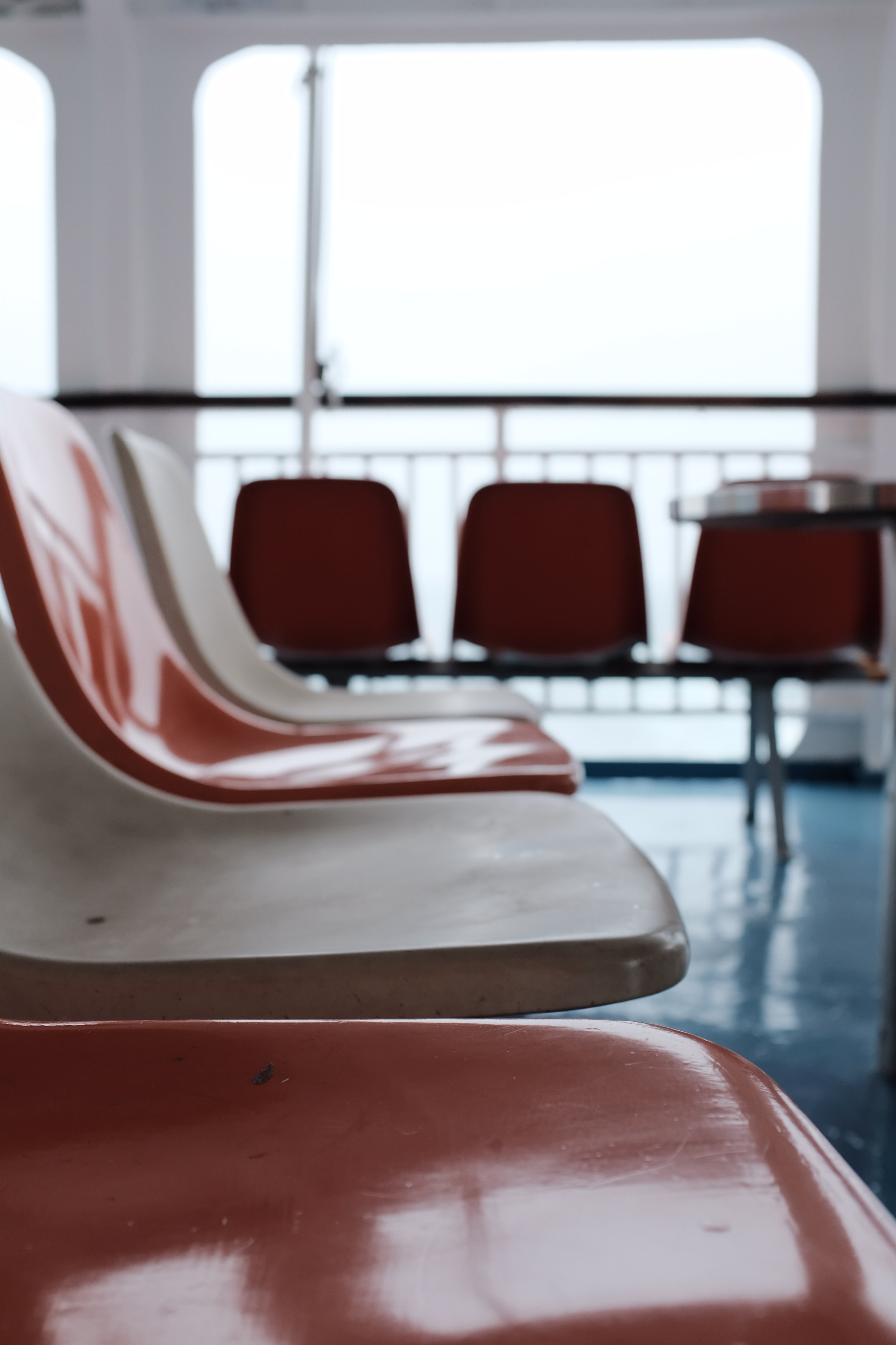 Rows of moulded plastic waiting-room chairs in shades of red and cream inside a bright indoor public space. The foreground focuses on the empty seats, while additional chairs and a small table appear softly blurred in the background near large windows. Natural light fills the space, creating reflections on the polished floor and chair surfaces.