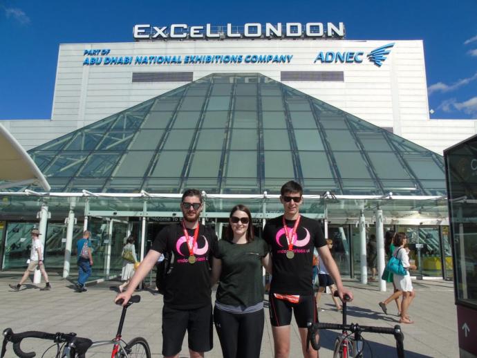 Three people stand outside the ExCeL London exhibition centre on a sunny day, two men flanking a woman in the middle. The two men wear matching black event T-shirts with pink logos and medals around their necks, and each stands beside a bicycle, while the woman wears dark athletic wear. Behind them, the building's glass pyramid entrance and "ExCeL LONDON" signage are visible, with other pedestrians passing by.