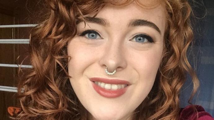 A close-up selfie of a young woman with curly red hair, blue eyes, a septum piercing, and red lipstick, smiling at the camera.
