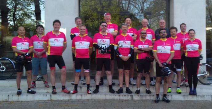 A group photo of about 15 cyclists, mostly men and one woman, standing together in matching pink, red, and yellow cycling jerseys, some holding helmets, posing outdoors in front of a building with trees in the background.