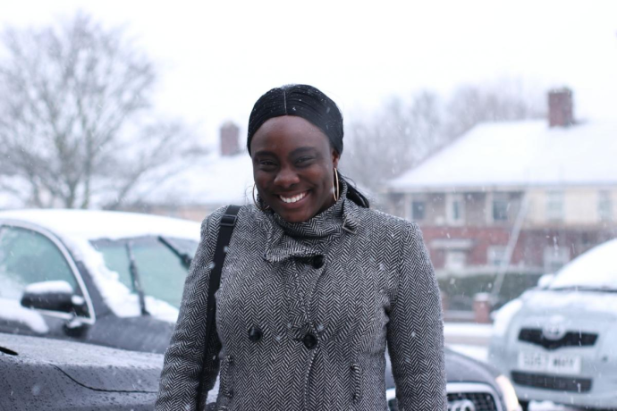 A smiling woman wearing a herringbone coat and hoop earrings stands outdoors in falling snow, with snow-covered cars and houses visible in the background.