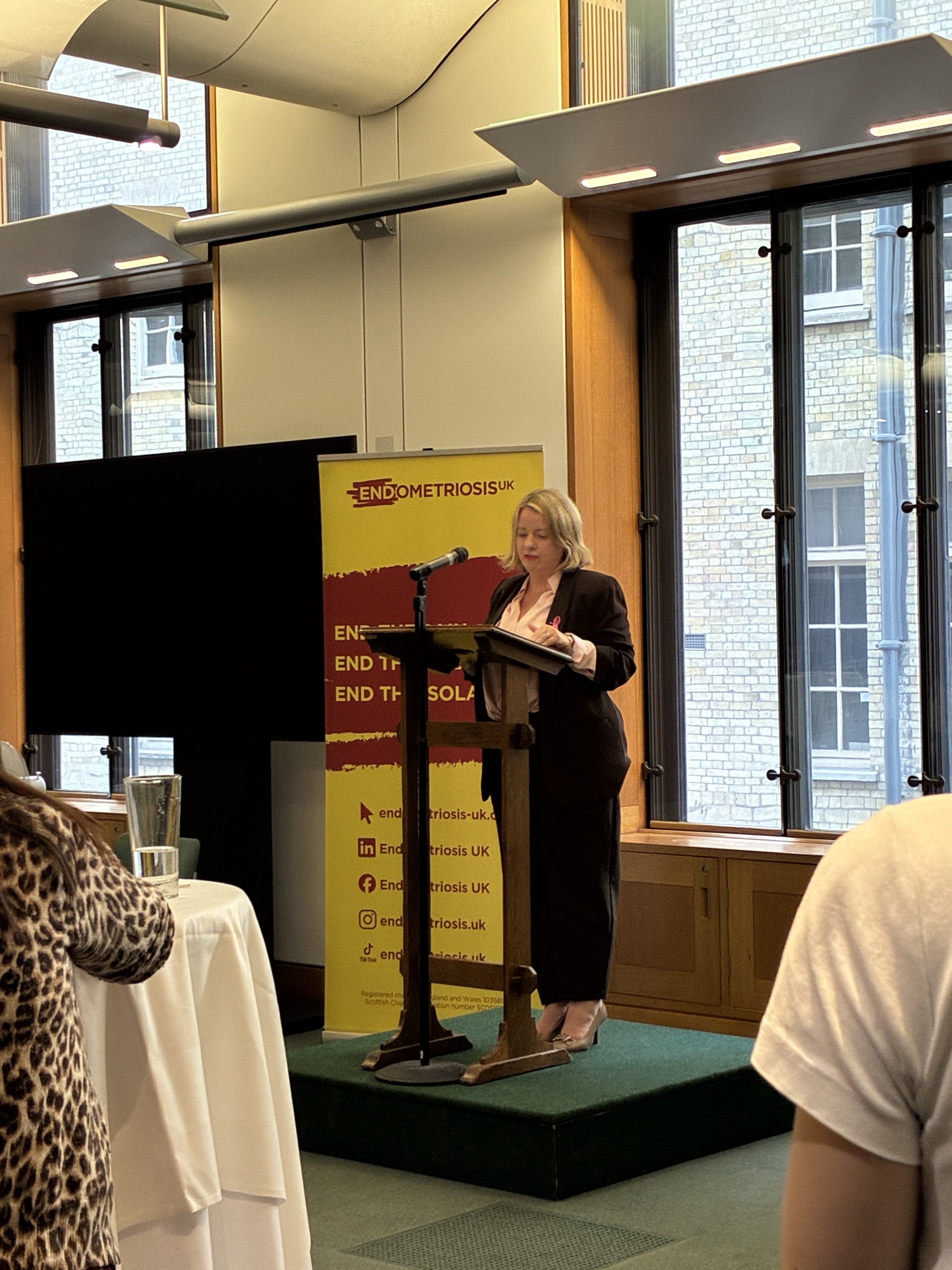 Kirsteen Sullivan MP stands at a lectern delivering a speech at an Endometriosis UK event. Behind the lectern is a yellow and red Endometriosis UK banner, with large windows and a brick building visible in the background. Audience members are seated in the foreground, facing the speaker.