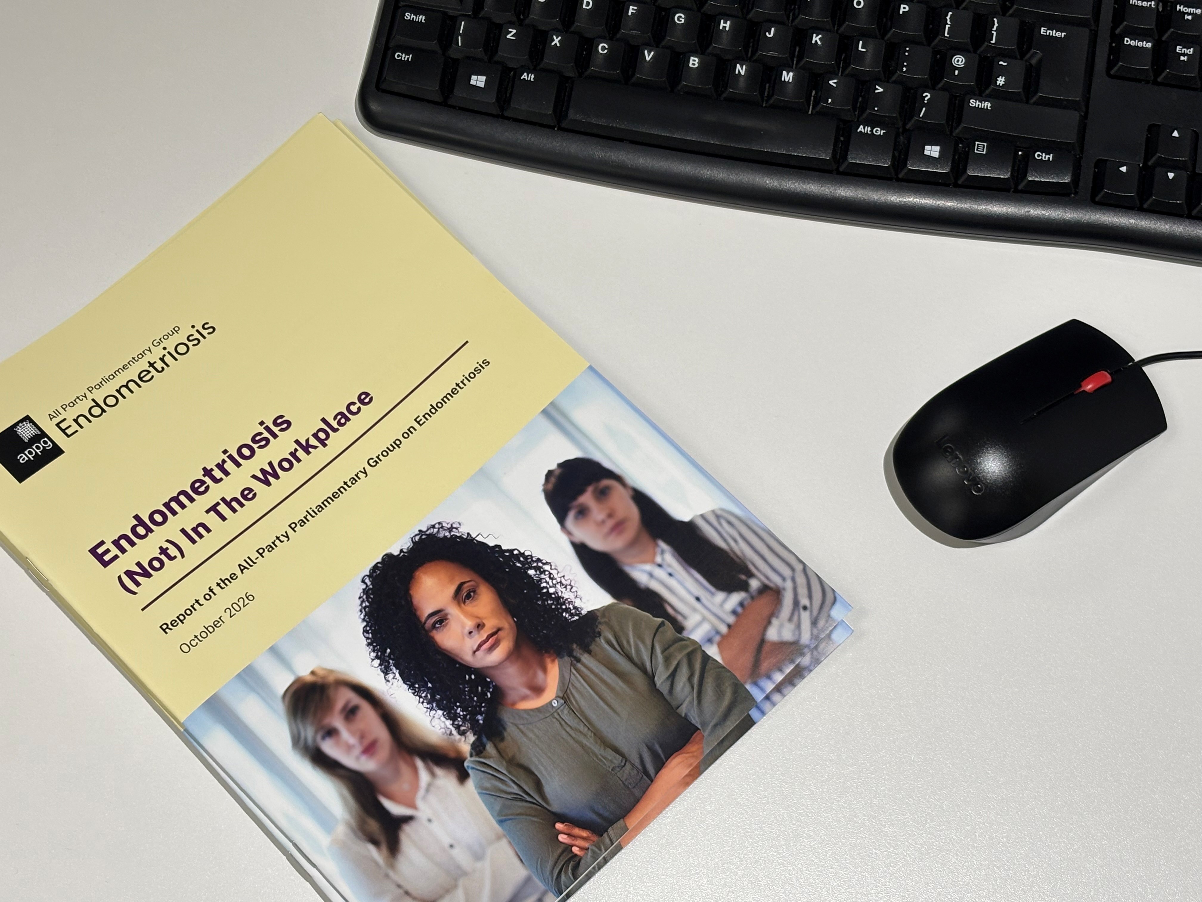 A copy of the All-Party Parliamentary Group on Endometriosis report, “Endometriosis (Not) In The Workplace” (October 2026), lies on a desk beside a computer keyboard and mouse. The report cover features a workplace image of three office workers, with the title prominently displayed on a pale yellow background.