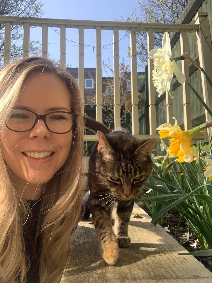 A photo of Helen smiling with her cat next to some yellow daffodils