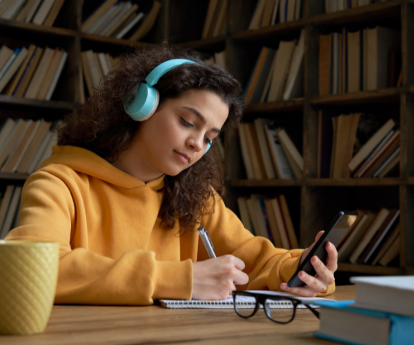 A teenage girl wearing a yellow hoodie and blue headphones studying in a library 