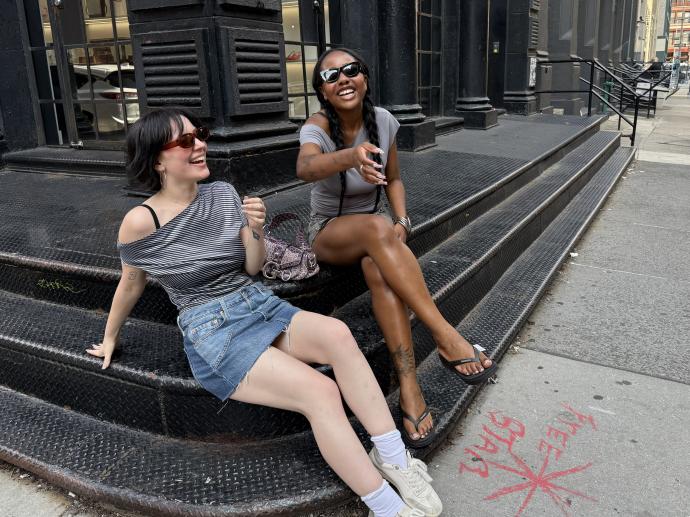 Two women laughing together while sitting on the steel steps of a building with dark storefronts. One woman has short dark hair, tinted sunglasses, and a striped off-shoulder top with a denim skirt; the other has long braids, black sunglasses, and a gray top with shorts. Red spray-painted text is visible on the sidewalk nearby.