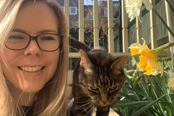 A photo of Helen smiling with her cat next to some yellow daffodils