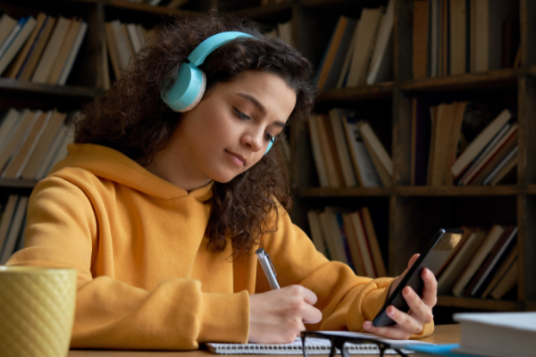 A teenage girl wearing a yellow hoodie and blue headphones studying in a library 