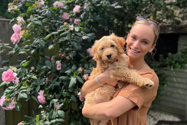 A photograph of Tannagh smiling, standing next to a rose bush and holding a puppy