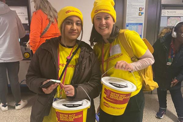 A picture of two of our Bucket Collection volunteers dressed in bright yellow Endometriosis UK hats and t-shirts, holding a yellow bucket each