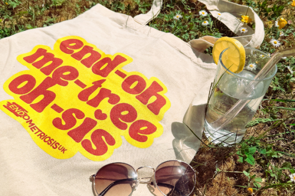 A cream tote bag printed with the phonetic spelling of 'endometriosis' in bold red retro text on a yellow background, reading 'end-oh me-tree oh-sis', with the Endometriosis UK logo. The bag is laid on grass in sunlight alongside a glass of water with a lemon slice, round sunglasses, and small wildflowers.