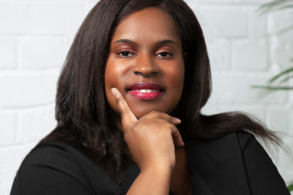 Professional headshot of Sanchia, a woman with long dark hair, smiling at the camera with her chin resting on her hand. She wears a black top and a silver watch, seated against a white brick wall with a plant in the background.