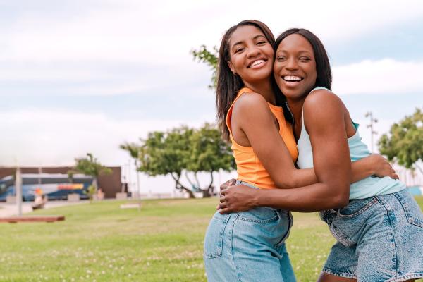 Two people standing on a grassy field outdoors with trees and an overcast sky in the background. The pair are embracing and facing the camera, wearing sleeveless tops and denim shorts. The open park setting includes a lawn stretching across the foreground, with scattered trees, a pathway and distant buildings visible in the background.